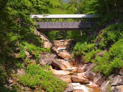 [A covered wooden bridge at least 30 feet above the boulders lining the path of the water heading downstream. Green vegetation lines both hillsides.]